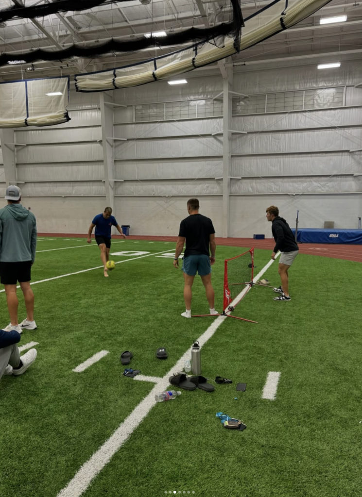 Four people, part of FCA (Fellowship of Christian Athletes), play a game with a small net on indoor turf; one kicks a ball as others watch. Sports equipment and personal items are scattered on the ground.